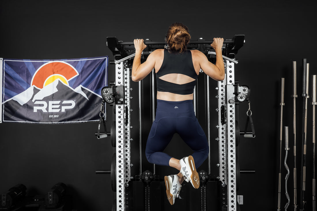 Person performing pull-ups on a pull-up bar (Ares 2.0 4-Post Pull-Up Bar + Pull-Up Bar Extension Brackets) with a REP Fitness banner in the background.