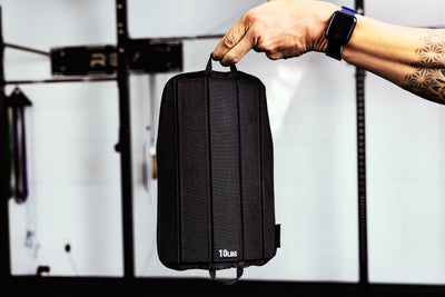 Person holding a black soft weight vest plate by loop  with '10Lus' branding against a blurred background