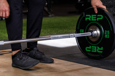 Close-up: Athlete secures a 25 lb REP Sport Bumper Plate 2.0 against a barbell collar, resting on the floor